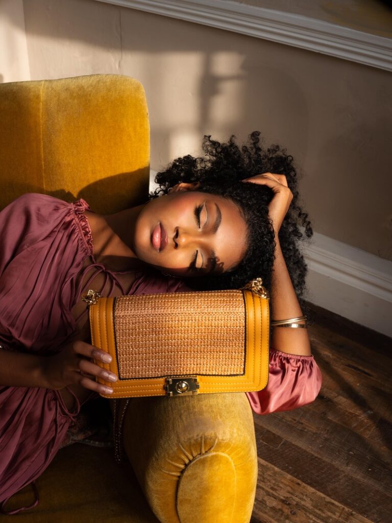 An image from the editorial campaign featuring the model Freselam. Freselam lounges on a yellow sofa chair with her eyes closed whilst holding the Cordelia in Havan Tan on the arm of the chair. 