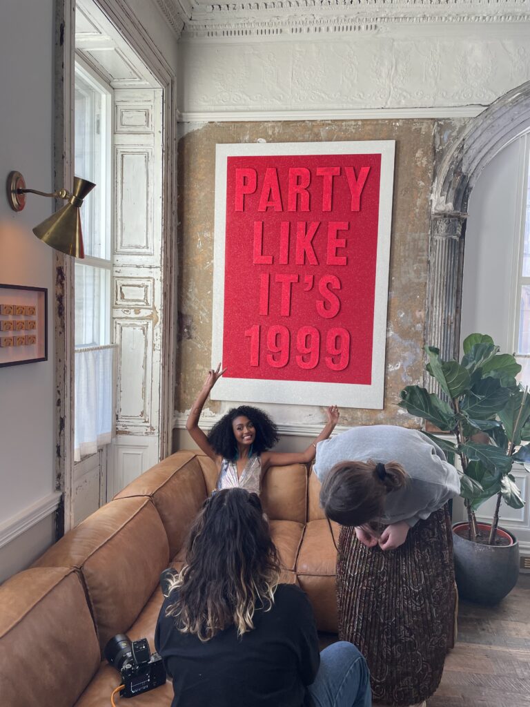 The model Freselam is captured behind the scenes between shots having fun with her hands in the air doing the peace sign. She is lounging on a long brown sofa with a 'Party like it's 1999' image on the wall behind. 